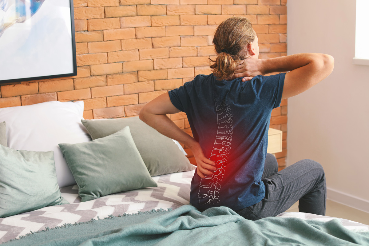 A person sitting on a bed experiencing neck and lower back pain in El Paso.